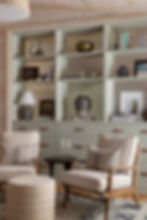 A cozy reading nook featuring plush armchairs and a soft ottoman, set against a backdrop of shelves displaying eclectic pottery, books, and artwork in a softly-lit living room.