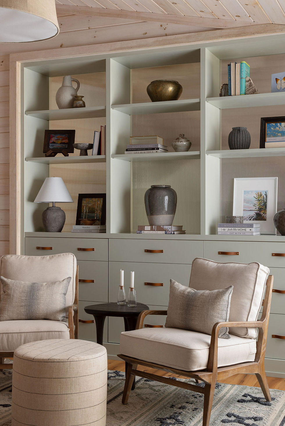 A cozy reading nook featuring plush armchairs and a soft ottoman, set against a backdrop of shelves displaying eclectic pottery, books, and artwork in a softly-lit living room.