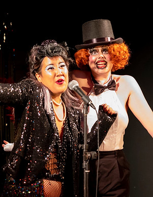 Two cabaret performers gather around a microphone singing. On the left there is a small brown Asian cabaret performer with sparkly makeup a lace flower in their dark hair and red lipstick, wearing fishnets a beaded leotard and a black sequin jacket. On the right is a slim white drag queen with cartoony makeup, a curly ginger wig and a top hat, wearing a white leotard with a bowtie and black trousers. They both look like they're having a jolly good time.