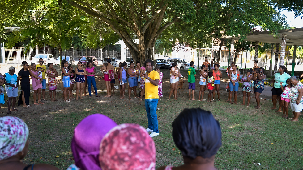 Homem de camiseta amarela fala a um grupo de mulheres reunidas sob uma grande árvore em parque, ao ar livre.