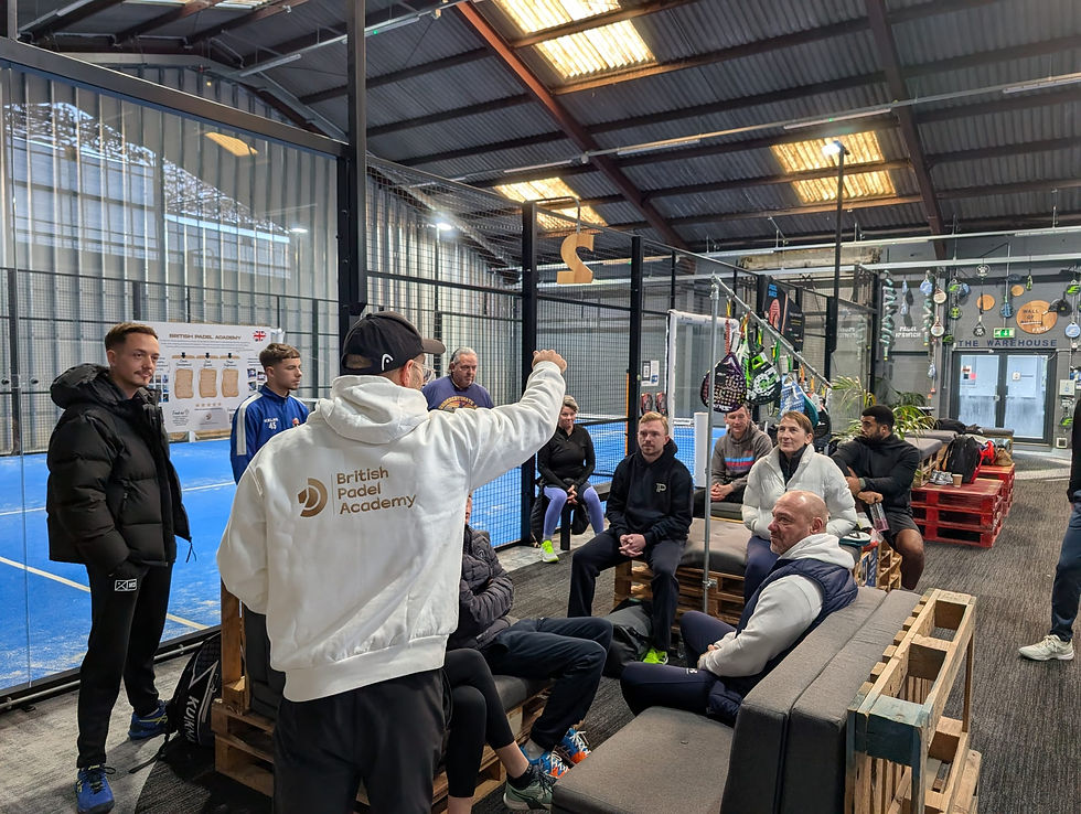 A group of people sit and stand in a padel court area, listening to Head Coach and founder of British Padel Academy Dan Troy