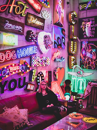 Girl poses for a portrait under a large wall covered in various neon signs.