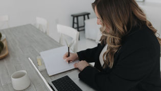 Woman in a black blazer writing in a notebook at a desk beside an open laptop, in a bright minimalist workspace.