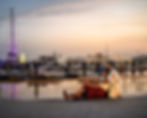 Couple sits on a bench at a marina during sunset, with boats and city skyline in the background. Warm, peaceful atmosphere couple photoshoot Dubai