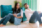 A man and woman sit back-to-back on the floor, smiling at each other. They're in a bright room with a teal sofa and green curtains couple photoshoot Dubai