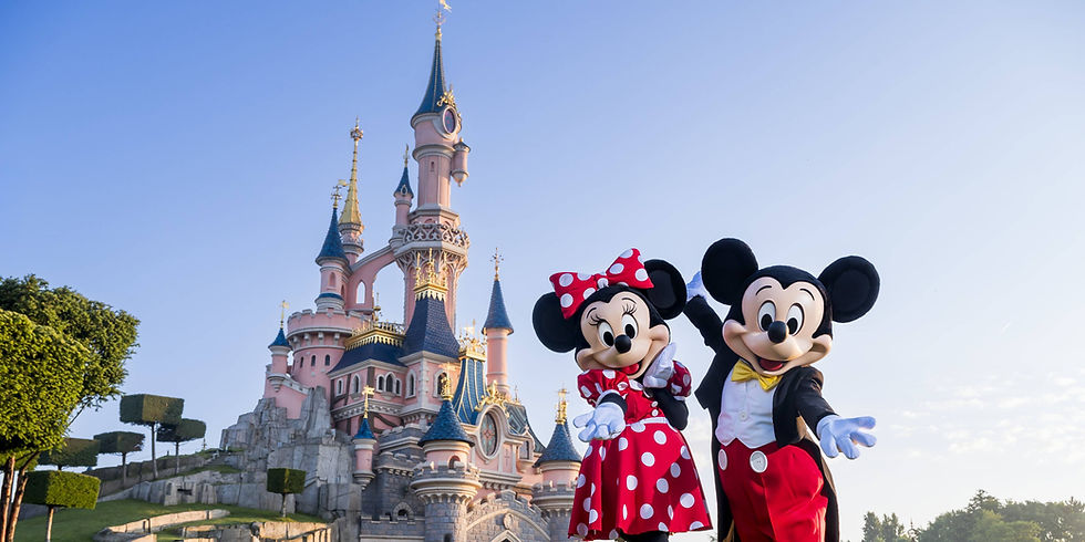 青空の下、ディズニーの城を背にミッキーとミニーが手を広げて笑顔で立つ、明るく楽しい雰囲気の写真