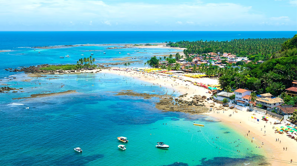 青く透明な海に囲まれたビーチ。人々が楽しそうに遊んでいる。ビーチにはカラフルなパラソル、多数のボートが浮かぶ。背景に緑豊かな森。