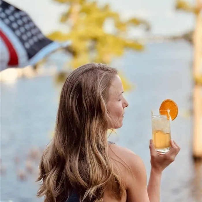 A woman holding a refreshing Bourbon Water cocktail poured over a glass of ice with a fresh orange slice garnish during the summer.