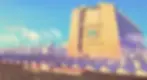 Futuristic cityscape with a large building displaying an "M" logo, surrounded by smokestacks and domed structures. Clear blue sky.