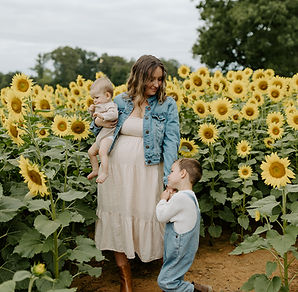 a mother holding her baby boy with her right hand and holding her 3 year old sons hand on her left hand standing in sunflower