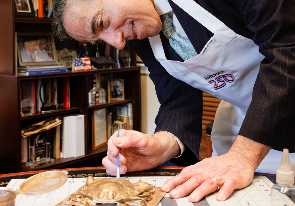 Maryland Representative Jamie Raskin wearing an America250 apron, carefully painting edible bronze paint onto a modeling chocolate crab sculpture in his Capitol Hill office.