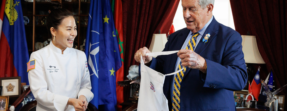 Rep. Joe Wilson holding up a commemorative America250 apron and laughing with cake artist Grace Pak, NATO and international flags visible behind them.