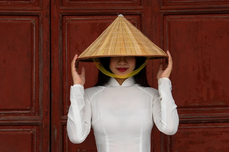 A person in a white outfit holds a conical hat, partially covering their face. Red wooden doors serve as the background.