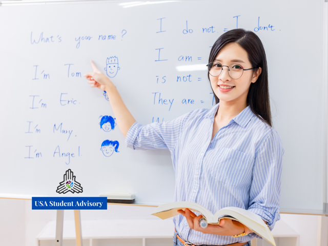 Smiling woman in glasses points at a whiteboard with English phrases. She's holding a book. Text: "USA Student Advisory."