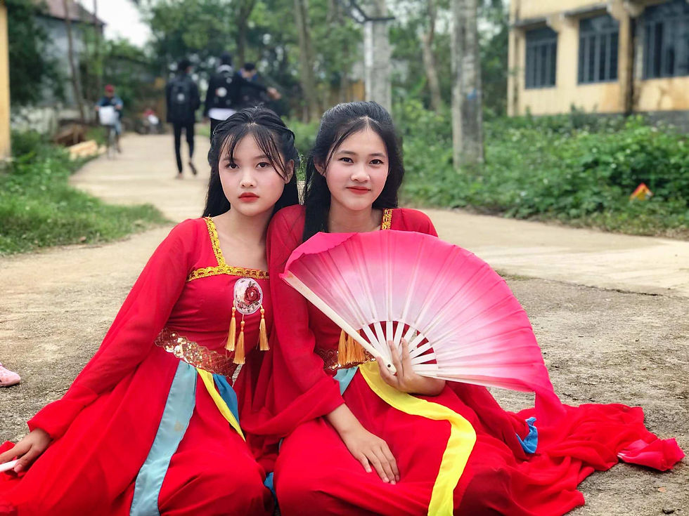 wo women in red dresses sit on a path holding a pink fan. Background shows greenery and buildings. They have a calm expression.