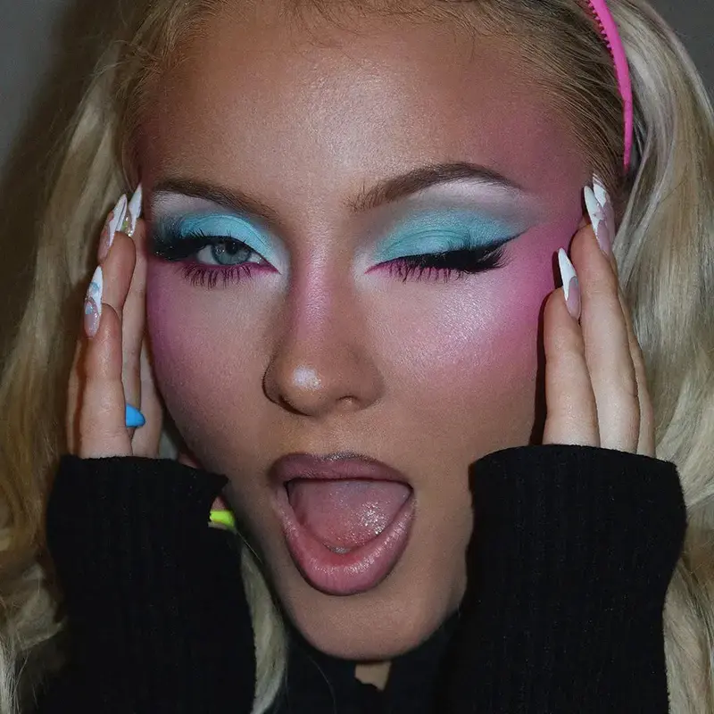 Close-up of a zara larsson winking with vivid blue-pink makeup, hands on her face, mouth open, wearing black sleeves and a pink headband.