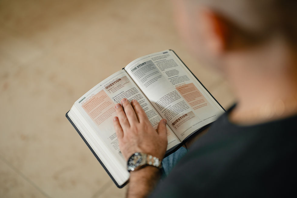 Open Bible viewed over the left shoulder of a man wearing a watch reading it using his hand as a guide/pointer