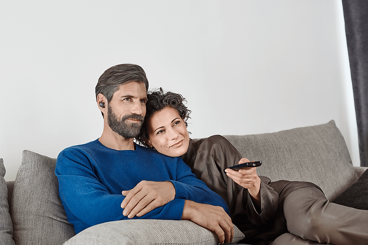 Auf dem Bild sitzt ein Paar entspannt auf dem Sofa. Der Mann trägt ein TV-Bluetooth Zubehör im Ohr, während die Frau eine Fernbedienung hält und sich an ihn lehnt.