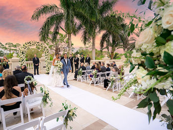 Bride and groom walk down a flower-adorned aisle surrounded by guests at a tropical wedding venue.