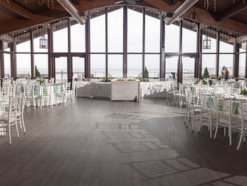 A wide-angle view of an indoor reception space with white chairs and large windows overlooking the ocean.