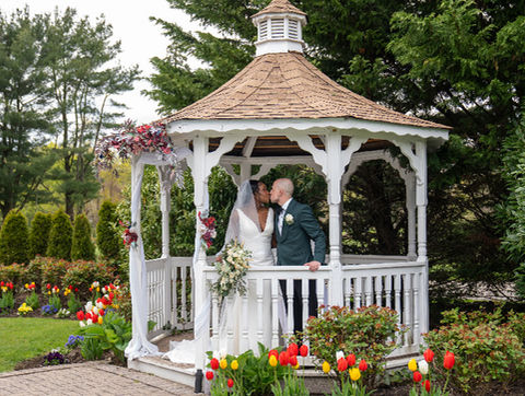 Bride and groom kissing inside a white gazebo decorated with flowers, surrounded by tulips and green landscaping.