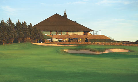 Golf clubhouse with a unique pyramid style roof, seen from the green with sand bunkers and trees under golden evening light.