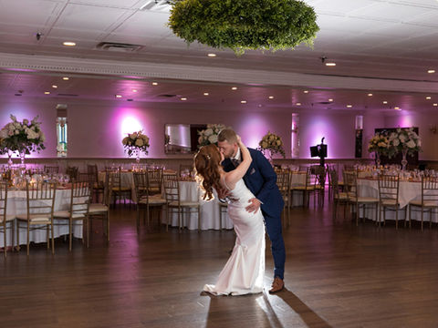 Groom dipping the bride for a dance in a decorated reception room with round tables, floral centerpieces, and purple uplighting.