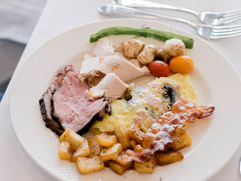 A breakfast plate with eggs, bacon, roasted potatoes, and sliced meat served on a white plate.