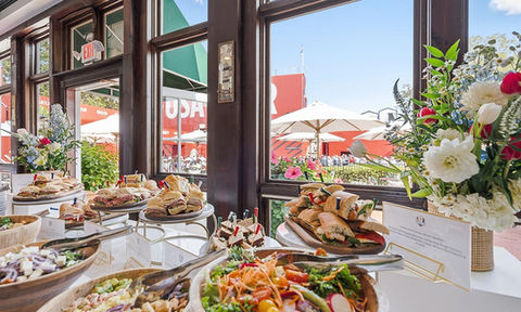 Assorted sandwiches and colorful salads displayed buffet-style near large windows, with floral arrangements and outdoor umbrellas visible outside.