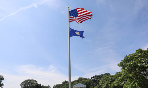 American flag and a second flag flying on a tall flagpole against a blue sky, with trees and part of a roofline below.