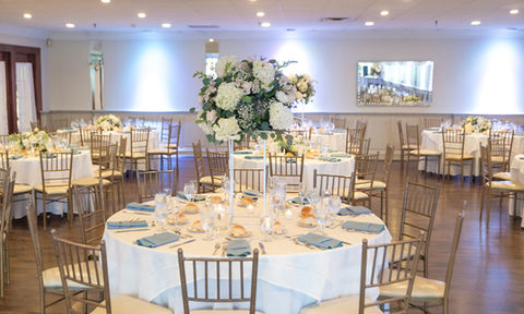 Reception table with tall floral centerpiece, blue napkins, place settings, and rolls on white plates, surrounded by gold chairs.