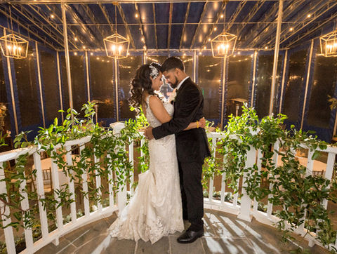 Bride and groom embracing under string lights on a decorated porch surrounded by greenery at night.