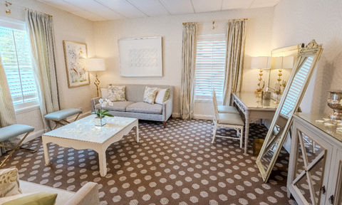 Bright lounge area with a vanity, leaning mirror, two small sofas, a coffee table with flower petals, and decorative lamps.