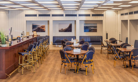 Casual dining area with wooden floors, bar seating, and navy blue chairs around wooden tables, decorated with coastal photography.