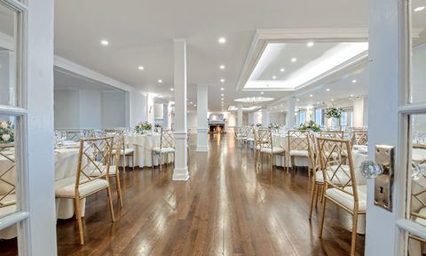 Bright, elegant banquet hall with round tables, gold chairs, hardwood floors, and white ceiling with recessed lighting.