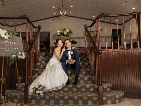 Bride and groom sitting on a carpeted staircase inside the venue beside a welcome sign with their names, floral decor, and lanterns.