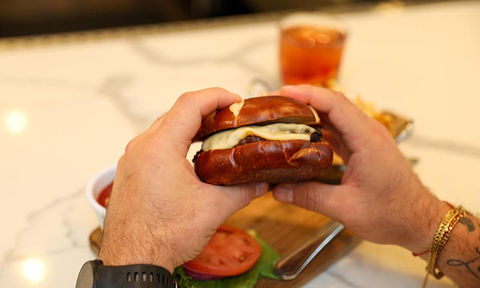 Hands holding a cheeseburger with a pretzel bun, with fries and a drink in the background.