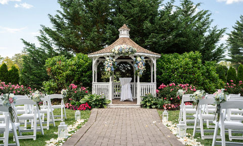Outdoor wedding ceremony setup with a white gazebo decorated in flowers, a brick walkway lined with flower petals and lanterns, and rows of white chairs on either side.