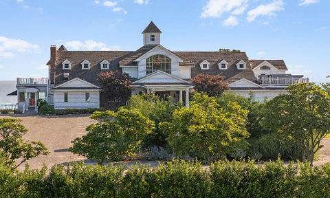 Close-up view of a large white waterfront house with dormer windows, a central cupola, and tree-lined landscaping.