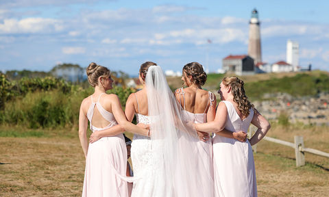 Bride standing arm-in-arm with her bridesmaids, all facing away toward a lighthouse in the distance on a sunny day.