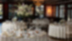 Elegantly set round wedding reception tables featuring tall white and blush floral centerpieces, gold chargers, and warm chandelier lighting.