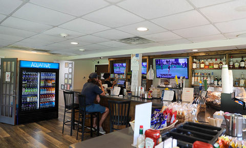Bar area with counter seating, a large refrigerator stocked with bottled drinks, liquor shelves, TV screens showing golf, and a snack counter with packaged food.