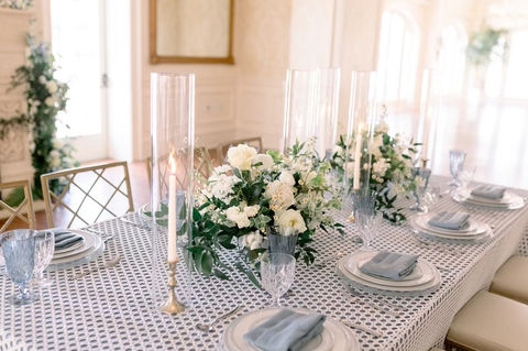 Elegant long reception table with blue and white floral centerpieces, tapered candles, and patterned linens in a bright hall.