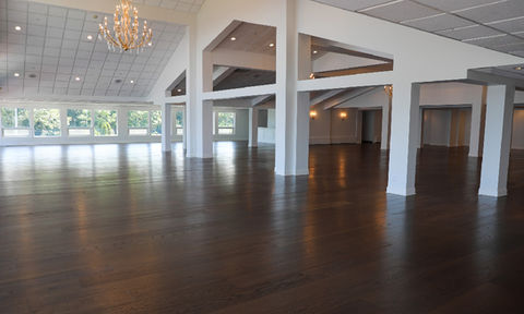 Large empty event space with polished wood floors, white columns, and chandeliers, featuring windows with tree views.