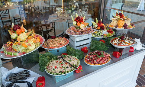 Buffet table with a variety of colorful appetizers and fruit displays, arranged with decorative vegetable carvings in front of a windowed dining area.