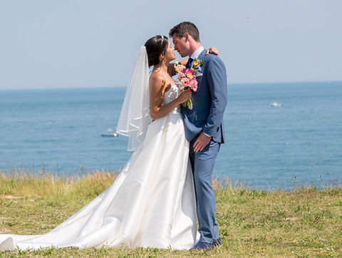Bride and groom standing together near the ocean, embracing on a grassy cliffside with boats visible in the water.