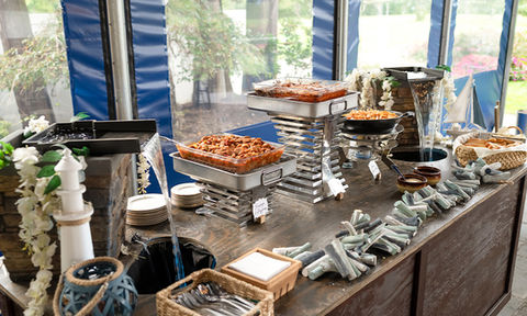 Decorative buffet station with flowing water features, metal trays of pasta and barbecue dishes, baskets of bread, and small signs labeling each item.