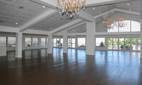 Spacious empty ballroom with chandeliers, vaulted ceiling, dark wood floors, and large windows overlooking greenery and a patio.