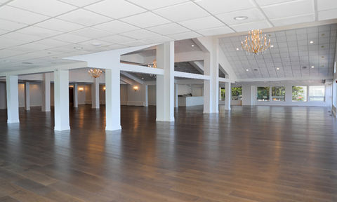 Empty banquet hall with white columns, wood floors, chandeliers, and multiple seating areas near large windows with outdoor views.