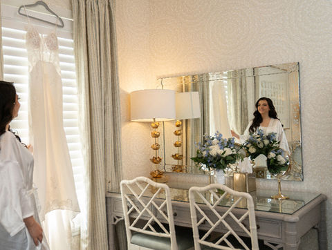 Bride in a white robe looking at her hanging wedding dress beside a mirror and floral arrangement on a vanity.